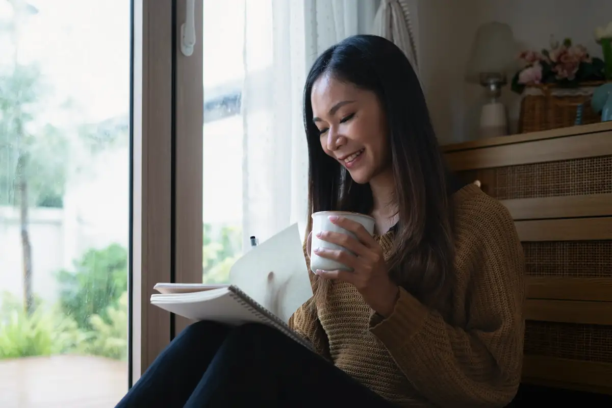 A smiling woman journaling by the window with a warm drink, representing reflection, healing, and processing pain through faith